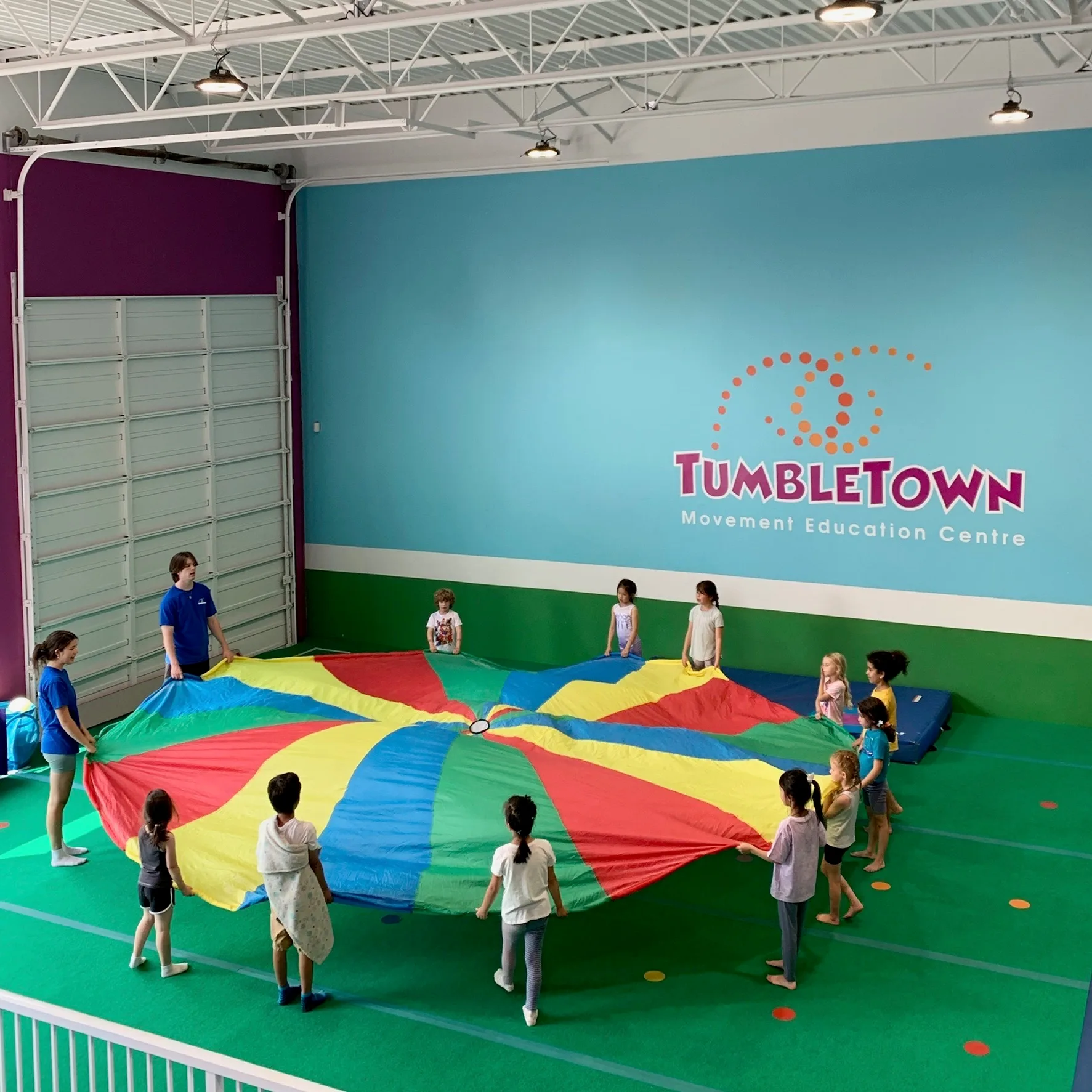 A class raising the rainbow parachute together under the TumbleTown wall logo