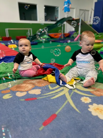 Two babies in TumbleTown shirts sitting on the rainbow parachute chasing bubbles