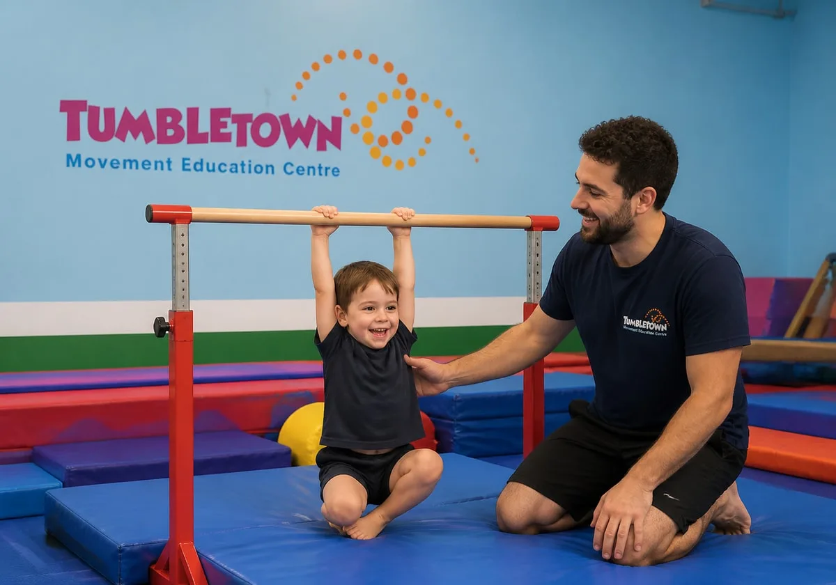 Four-year-old hanging from a low gymnastics bar while a male TumbleTown instructor spots