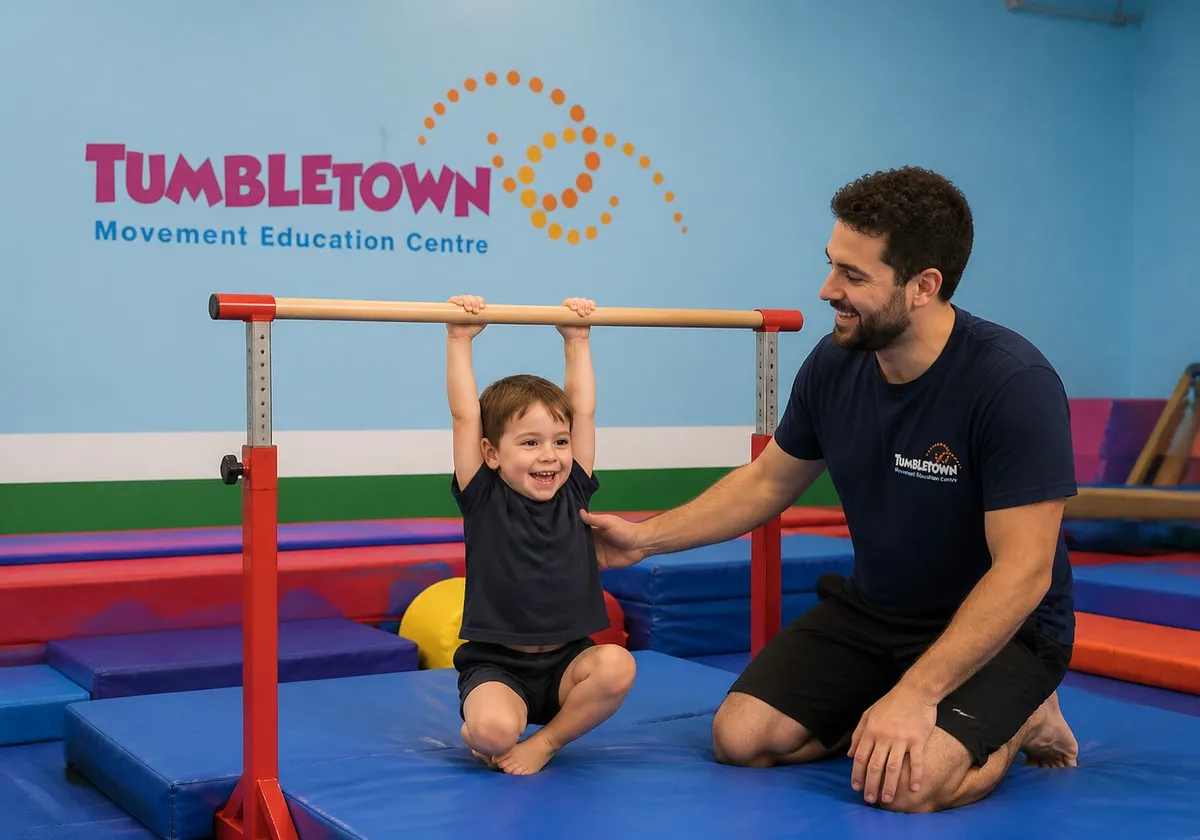 Four-year-old hanging from a low gymnastics bar while a male TumbleTown instructor spots