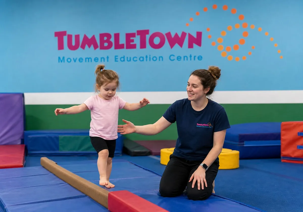 Three-year-old walking along a low padded beam with a female TumbleTown instructor alongside