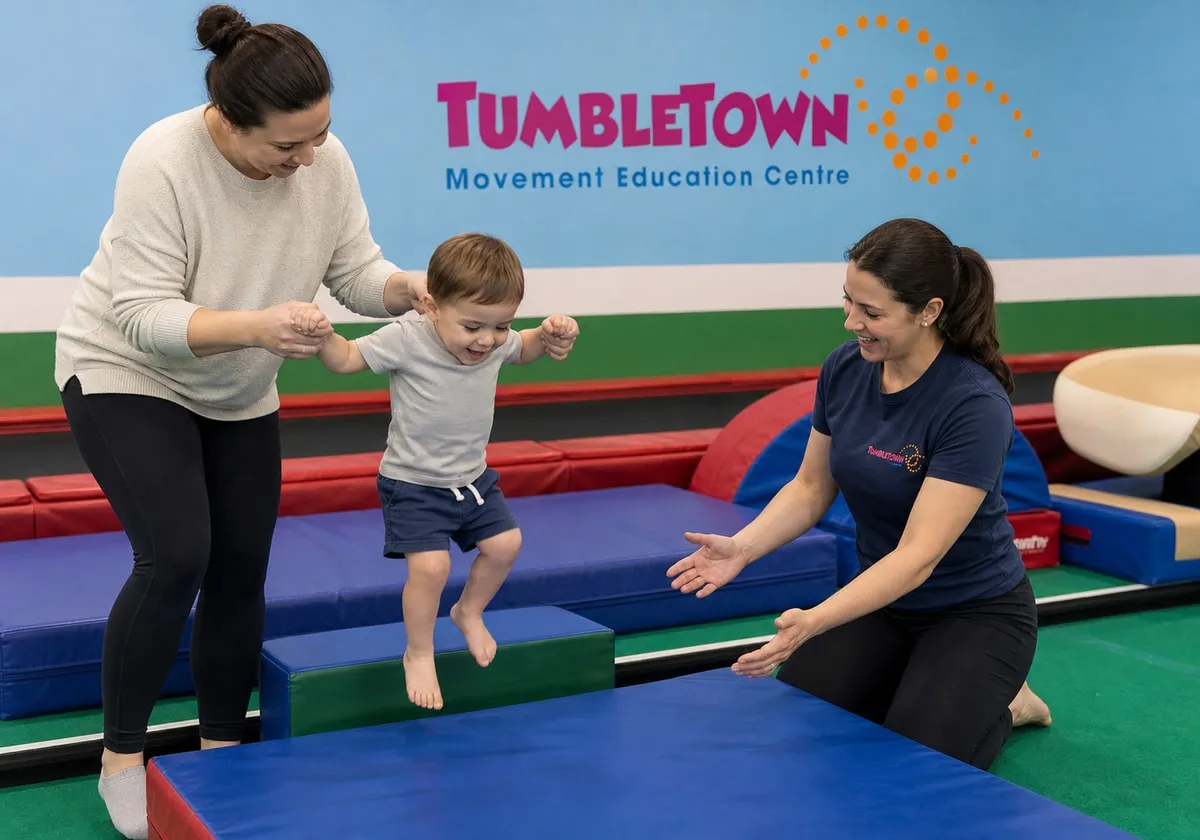 Two-year-old jumping from a low foam step while a parent holds their hands and a female instructor spots